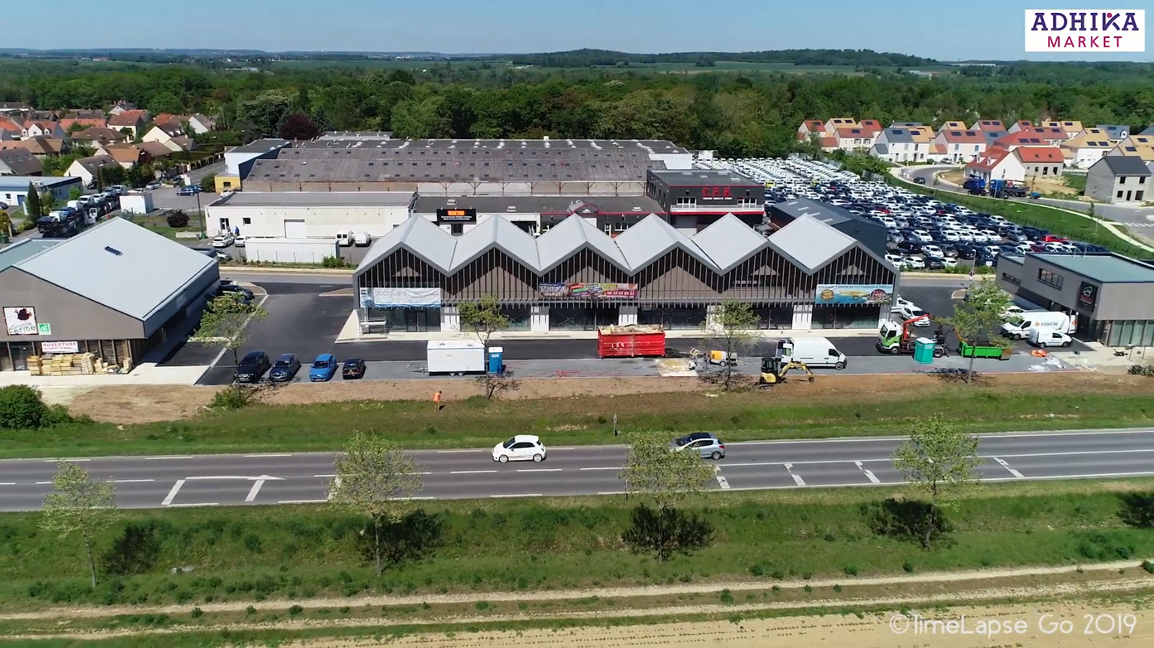 Vue aérienne du centre commercial Adhika Market avec des voitures garées devant.